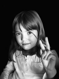 Portrait of cute girl showing peace sign while sitting against black background