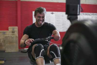 Confident male adaptive athlete exercising with rowing machine against wall in gym