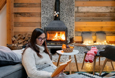 Young woman in warm sweater sitting by fireplace, reading a book.