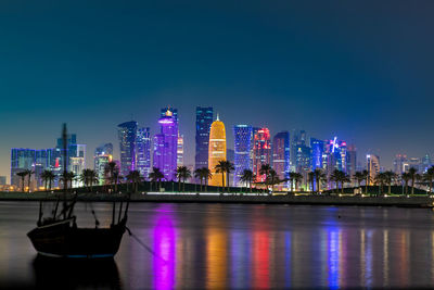 Illuminated buildings by river against blue sky at night