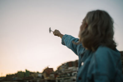 Side view of unrecognizable dreamy female sitting on roof of building and casting spell with magic wand in outstretched hand while looking away