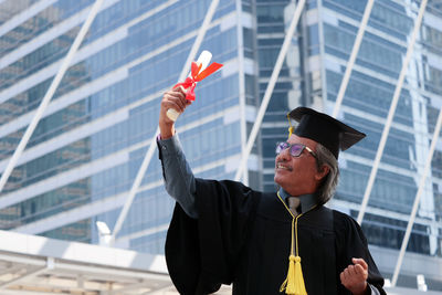 Businessman in graduation gown clenching fists against office building