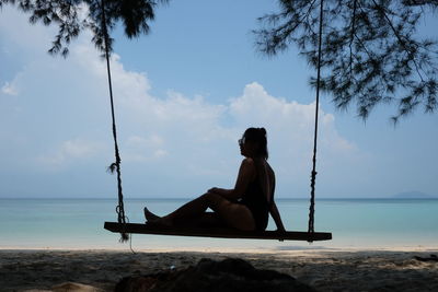 Silhouette woman sitting on swing at beach against sky