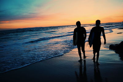 Rear view of silhouette friends standing at beach during sunset