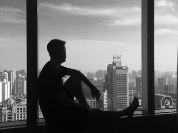 Full length of man sitting by window against sky in city