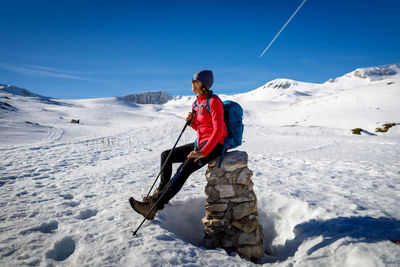 Woman rests and sunbathes sitting on a low wall, while hiking in the snow in the mountains. 