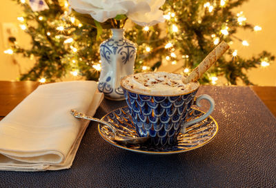 Cup of hot chocolate in a fancy bone china cup with gold and a silver spoon in front of a christmas