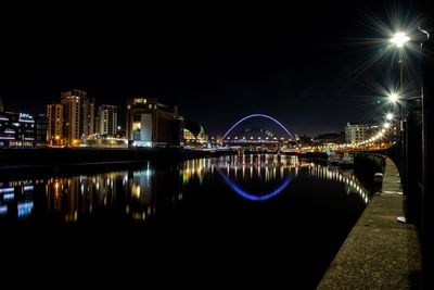 Illuminated bridge over river by buildings against sky at night