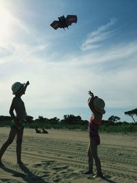 Full length of boy with sister flying kite at sandy beach against sky