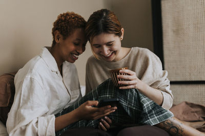 Happy non-binary couple watching smart phone while sitting on bed at home