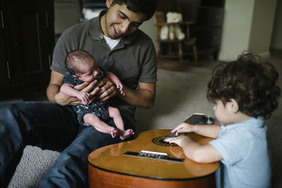 Baby boy playing guitar while sitting by father and newborn brother at home