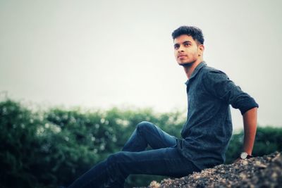 Thoughtful young man sitting on rock against clear sky