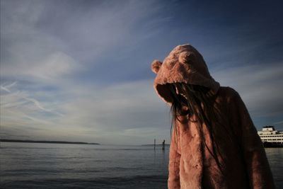 Young woman wearing hooded jacket while standing by sea against sky