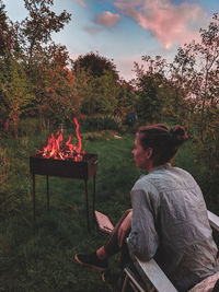 Rear view of woman sitting by campfire against sky