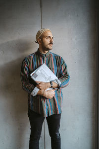 Thoughtful male adult student holding book looking away in front of gray wall at college