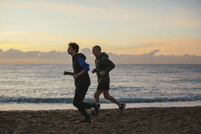 Side view of father and son jogging at beach against sky during sunset