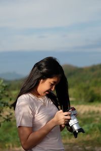 Young woman looking at dslr while standing on grassy field against sky