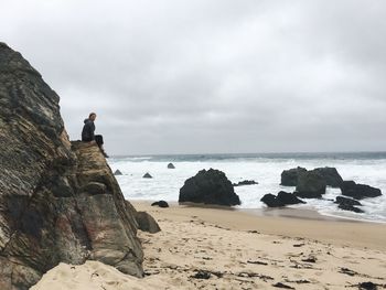 Woman sitting on rock mountain at beach against cloudy sky