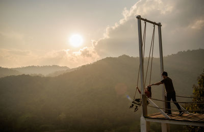 Woman sitting on swing while man pushing her against mountain and sky