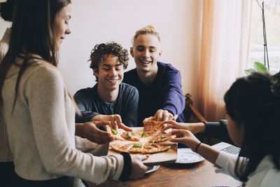 Teenage girl serving pizza to happy friends at dining table