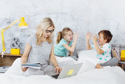 Siblings playing on bed while mother working over laptop at home