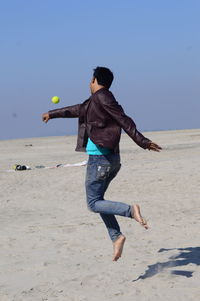 Man jumping while playing with tennis ball at beach against clear sky