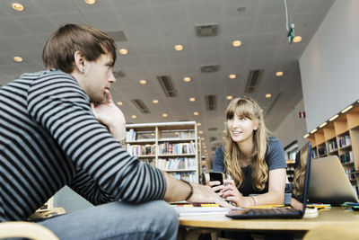 Young woman with male friend discussing at table in college library