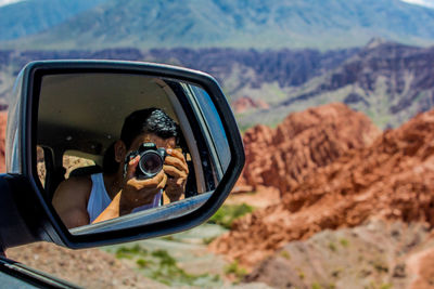 Man photographing while reflecting on side-view mirror of car