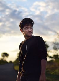 Young man wearing eyeglasses looking away while standing against sky during sunset