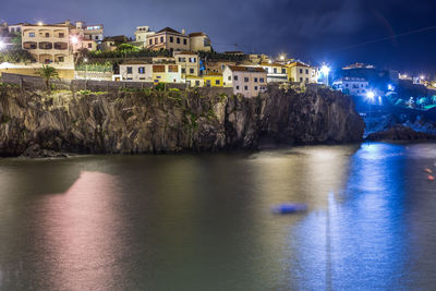 Illuminated buildings by sea against sky at night