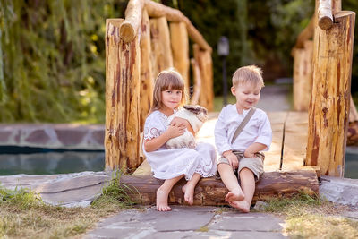 Portrait of girl carrying rabbit while sitting with brother on wooden bridge in park