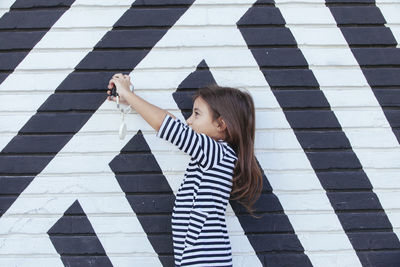 Side view of girl photographing with camera while standing against wall