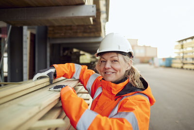 Portrait of smiling female worker in protective workwear stacking planks at industry