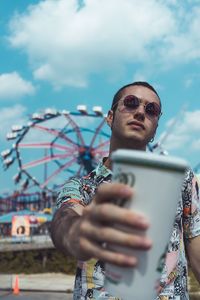 Portrait of young man wearing sunglasses holding coffee while standing against amusement park