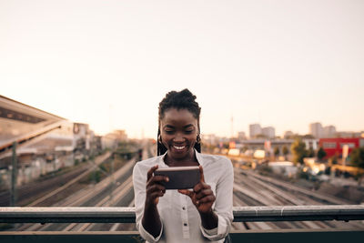 Young woman using phone while standing on city against sky