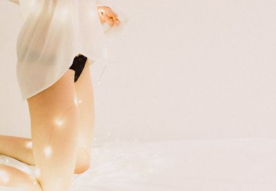 Midsection of young woman with illuminated string lights kneeling on bed against wall