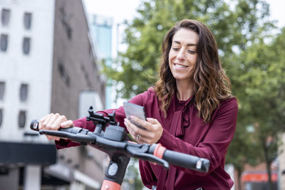 Smiling businesswoman using mobile phone to unlock electric bicycle in city