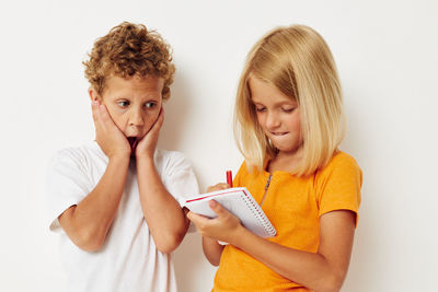 Cute girl writing in notebook while standing with brother against wall