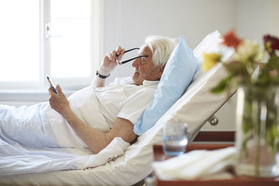 Side view of senior man wearing eyeglasses while using smart phone in hospital ward