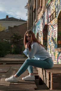 Portrait of confident young woman sitting on wood against wall in city