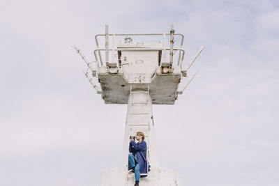 Low angle view of woman photographing while sitting on ladder at lookout tower against sky