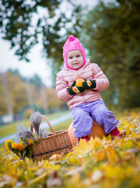 Portrait of cute girl holding orange while sitting on pumpkin over autumn leaves against trees at public park