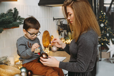 Mother and child on christmas kitchen paying with decor and baubles. woman with son prepare home for