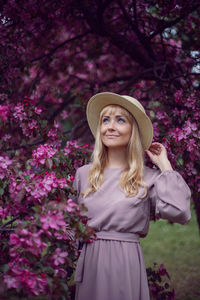 Portrait young beautiful woman in a pink dress and hat stands by a blooming pink apple tree