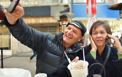 Man taking selfie while sitting with woman at sidewalk cafe