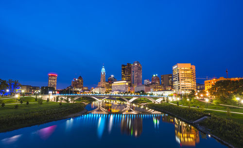 Illuminated buildings by river against blue sky
