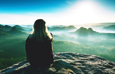 Fair hair girl with warm jacket sit on rocky mountain top against bright morning sky enjoy panorama