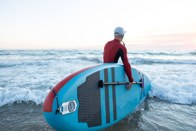 Back view of unrecognizable male surfer in wetsuit and hat carrying paddle board and entering water to surf on seashore