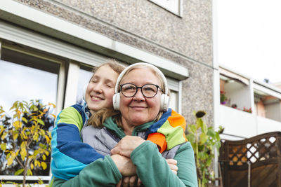 Girl embracing grandmother listening music through headphones outside house