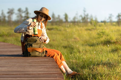 Woman botanist in hat sitting on wooden path through peat bog swamp, takes a thermos from a backpack